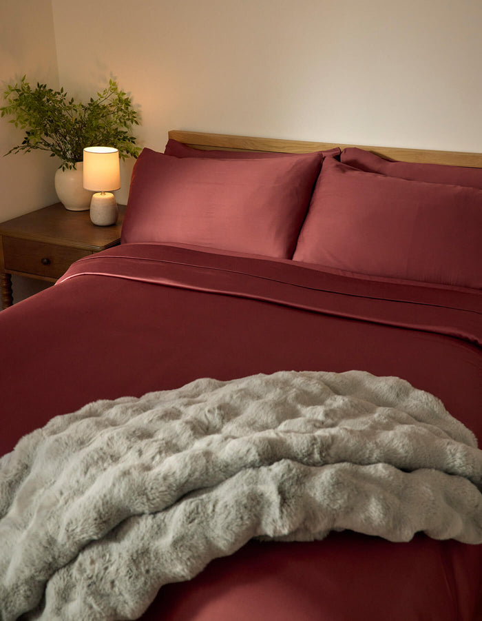 A neatly made bed featuring Cozy Earth’s Bamboo Sheet Set in deep red, paired with a light gray textured blanket, a wooden nightstand with a lamp and green plant, all set against a plain cream wall.|Color:Mulberry