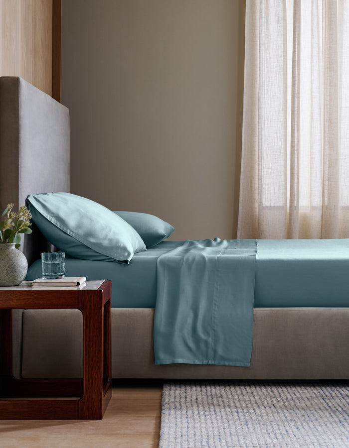 A neatly made bed in a minimalist bedroom setting, featuring a light blue Bamboo Sheet Set set against a neutral gray headboard and beige walls. |Color:River