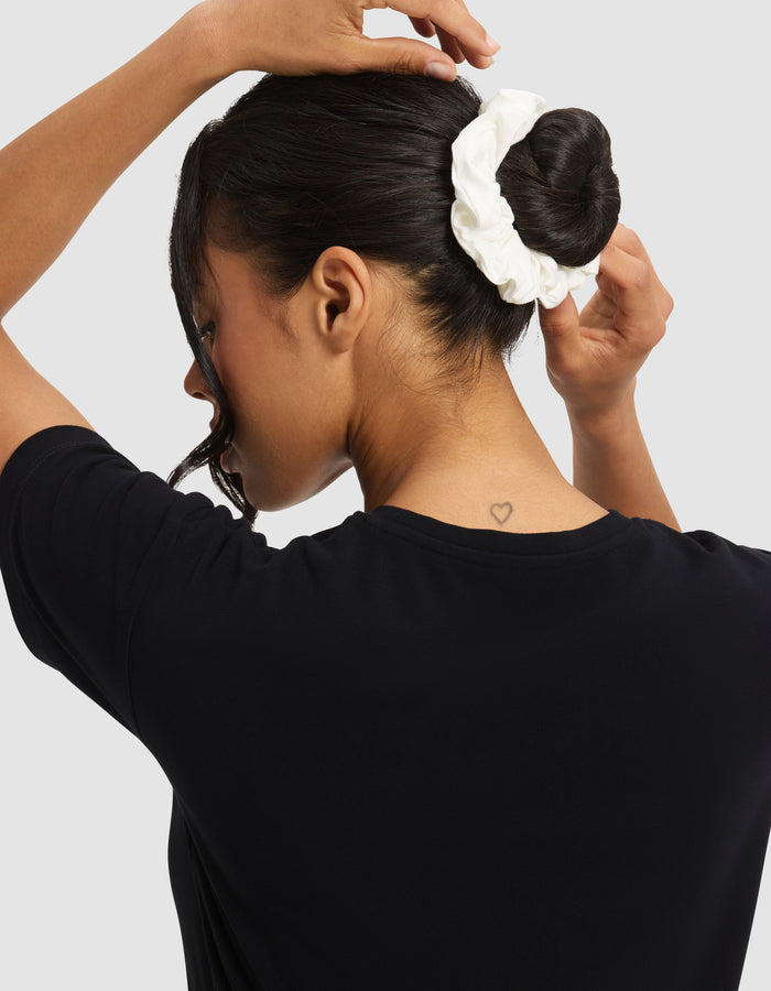 A person with dark hair ties their hair in a bun using a white Cozy Earth Bamboo Stretch-Knit Scrunchie. They wear a black shirt, have a small heart tattoo on the back of their neck, and stand against a light gray background.|Color:Black/Ivory/Grey