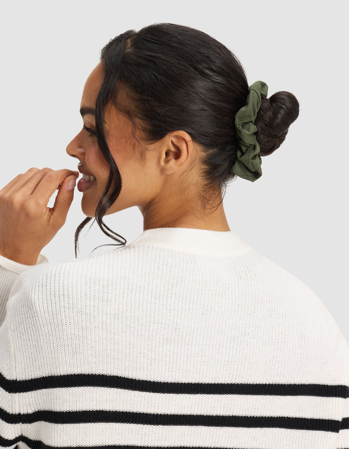 A woman with dark hair in a bun, secured by a Cozy Earth Bamboo Stretch-Knit Scrunchie in olive green, wears a white sweater with black stripes and smiles to the side against a plain background.|Color:Olive/Fern/Heather Grey