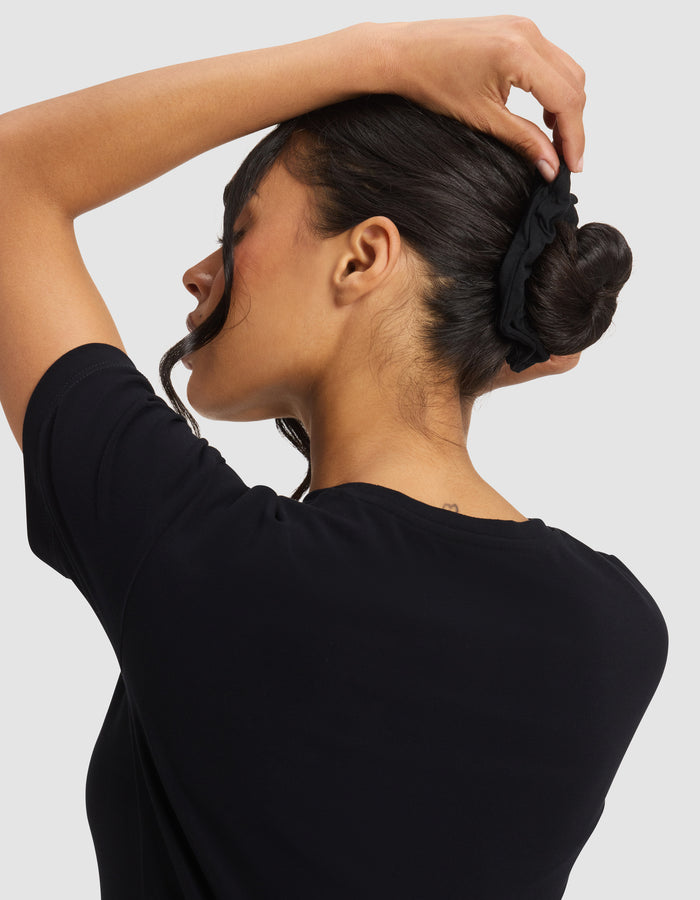 A woman with dark hair, wearing a black shirt, faces away and adjusts her bun with a Cozy Earth Bamboo Stretch-Knit Scrunchie. A loose strand falls by her face.|Color:Black/Ivory/Grey