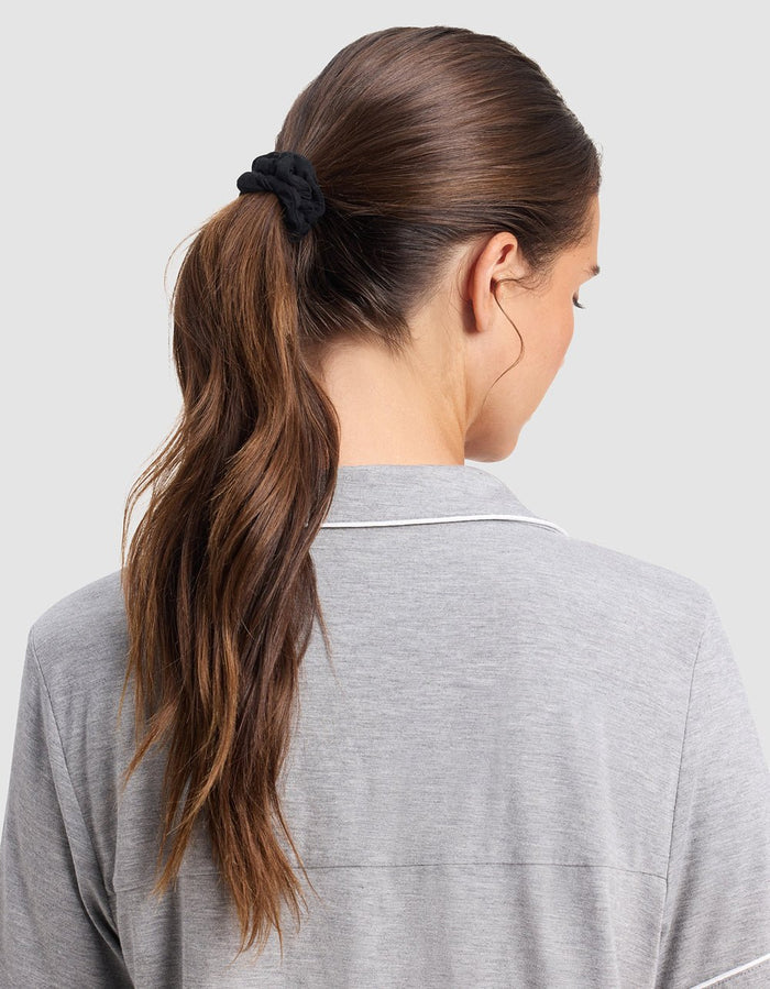 A woman with long brown hair tied in a ponytail with a black Cozy Earth Bamboo Stretch Knit Skinny Scrunchie, wearing a light gray collared shirt, is seen from the back against a plain light background.|Color:Black/Ivory/Light Grey