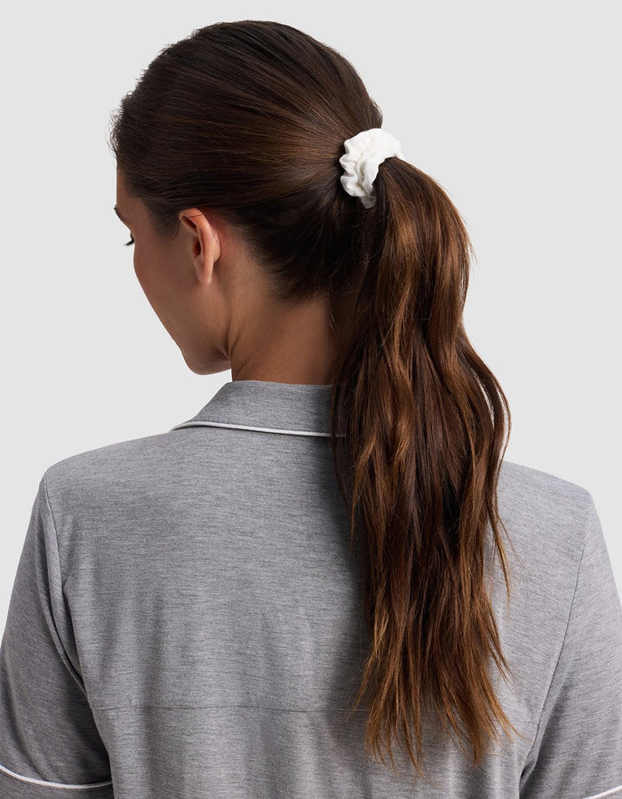 A woman with long brown hair in a ponytail secured by a white Cozy Earth Bamboo Stretch Knit Skinny Scrunchie, wearing a grey collared shirt, is shown from behind against a plain light background.|Color:Black/Ivory/Light Grey
