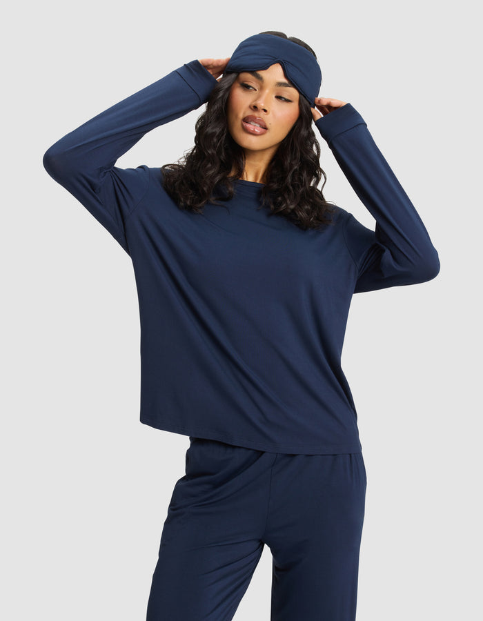 A woman wearing a navy blue pajama set poses with her hands touching the sides of a Cozy Earth Bamboo Stretch-Knit Sleep Mask, standing against a plain light background.|Color:Navy