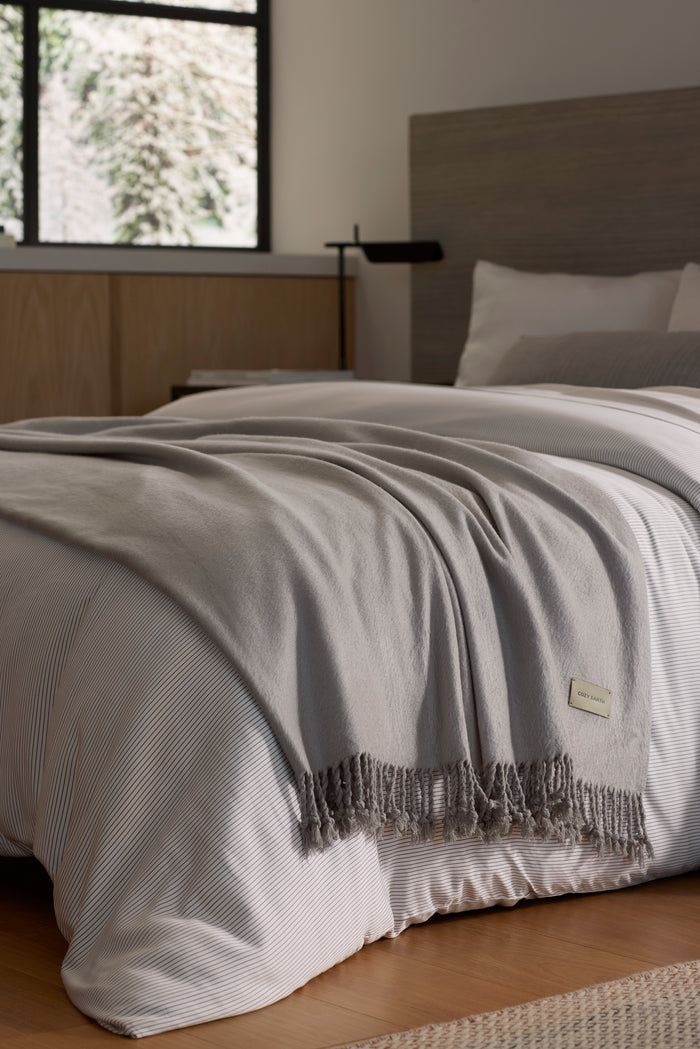 A neatly made bed with white and light gray bedding is topped with the Cozy Earth Bamboo Tassel Throw in gray. The room features a wooden headboard, light wood furniture, and a window letting in natural light.|Color:Dove Grey