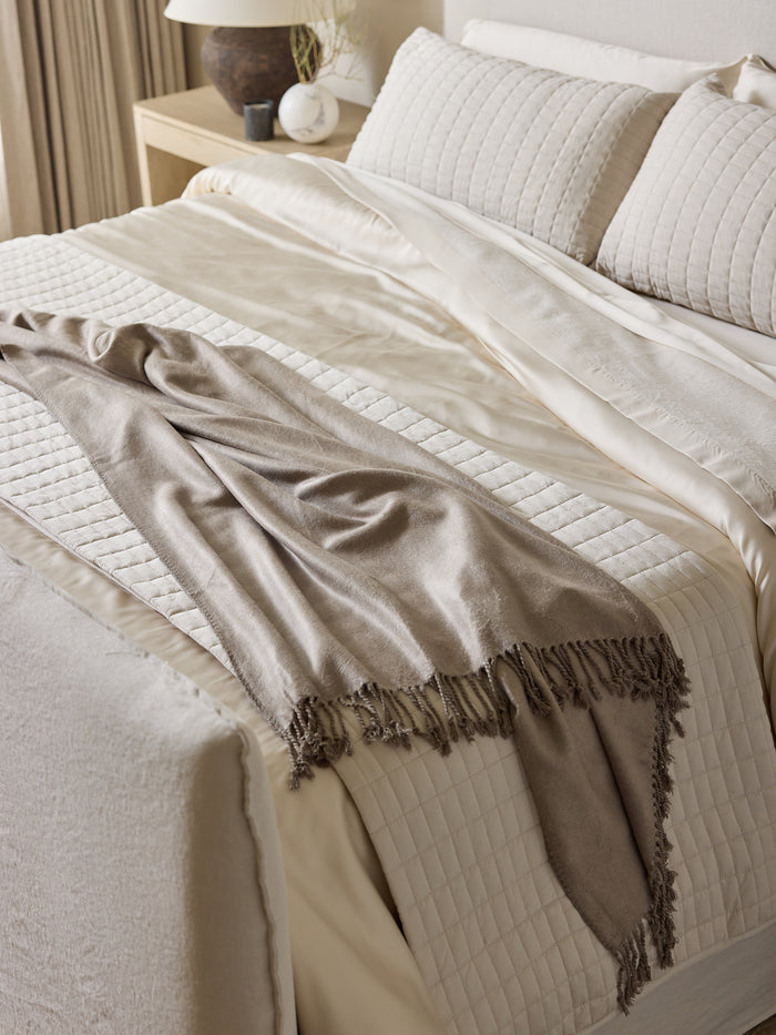 A neatly made bed with cream-colored sheets, a quilted blanket, and two pillows. The Bamboo Tassel Throw from Cozy Earth, featuring fringed edges, is casually draped across the lower part of the bed. The bedside table holds a lamp, a small plant, and some decorative items. |Color:Walnut