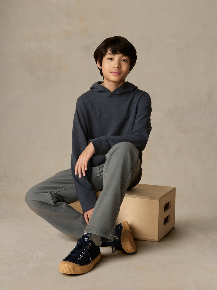 Child sitting on a wooden box wearing a blue hoodie with grey pants against a beige background