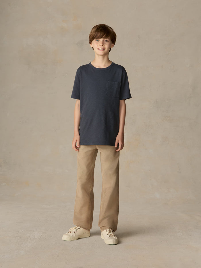 Child wearing a blue pocket tee and beige pants against a beige background