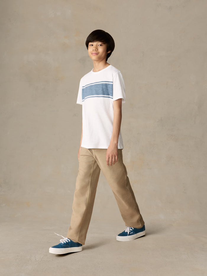 Child wearing a white shirt with blue stripe and beige pants against a beige background