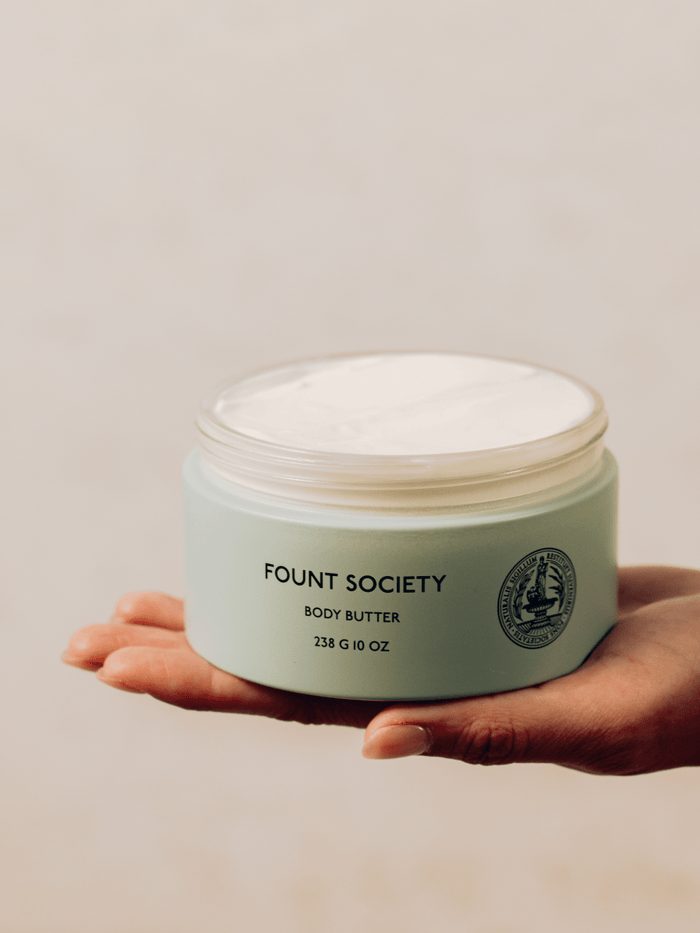 A hand holding a light green container of Cozy Earth Body Butter with a slightly opened lid, revealing the creamy product inside. The container has a 238 g (10 oz) label and a circular emblem. The background is softly blurred.|Size:Full Size