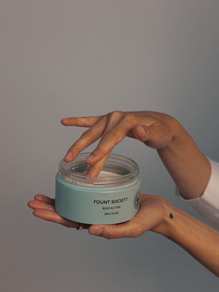A person dips their fingers into an open jar of "Cozy Earth" body butter, held in their other hand. The jar is teal with black text, and the person's hands are positioned against a neutral background. A small tattoo is visible on their wrist.|Size:Full Size