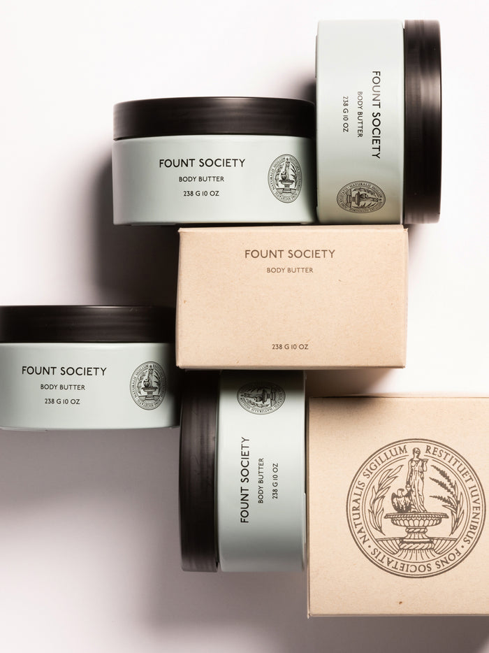 The image features an arrangement of body butter containers from the brand "Cozy Earth." The containers, labeled "Body Butter" with a quantity of 236 g (8 oz), are stacked and include a mix of uncovered and boxed items, showcasing the product branding and logo.|Size:Full Size