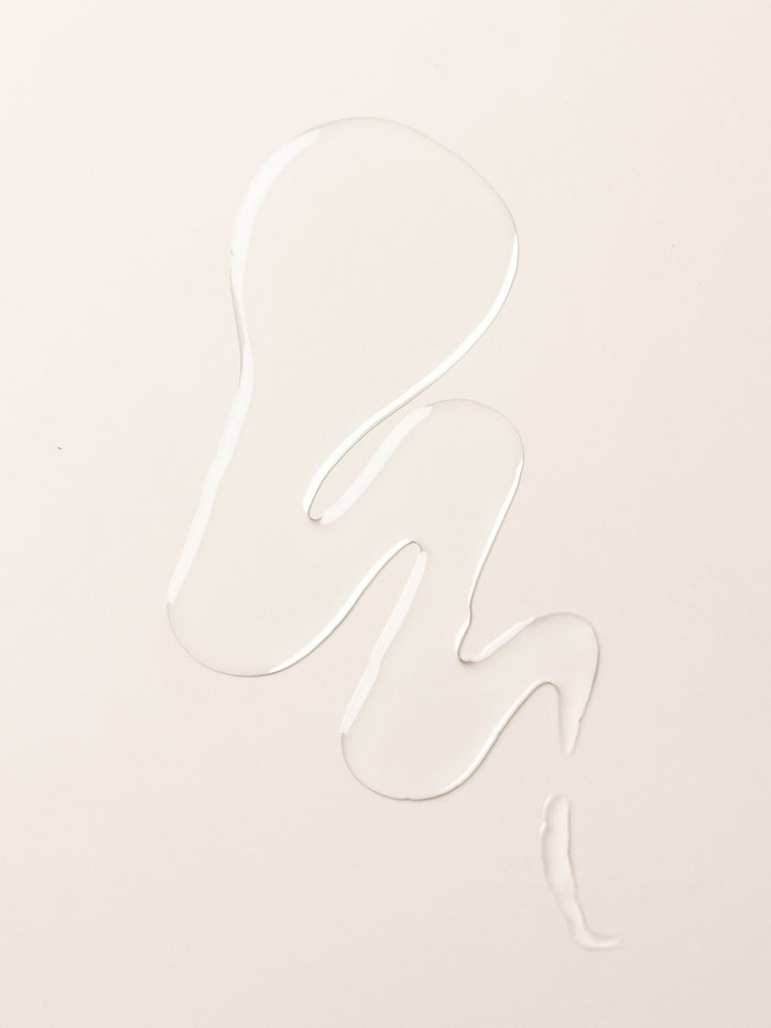 A clear liquid body oil from Cozy Earth forms an abstract, wavy shape on a smooth, off-white surface.