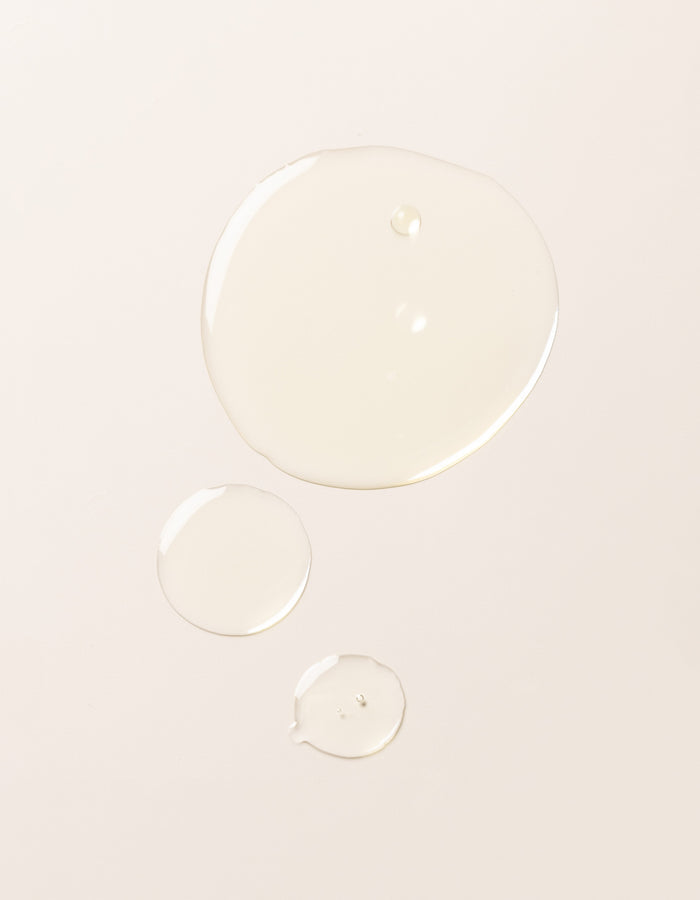 Three droplets of Cozy Earth's Brightening Vitamin C Oil, each of a different size, are spaced apart on a smooth, light-colored surface.