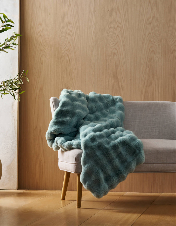 A light gray chair with wooden legs displays the Bubble Cuddle Blanket by Cozy Earth in textured teal, set in a room with light wood-paneled walls, a potted plant, and a white canvas. Natural light brightens the space.|Color:Tide