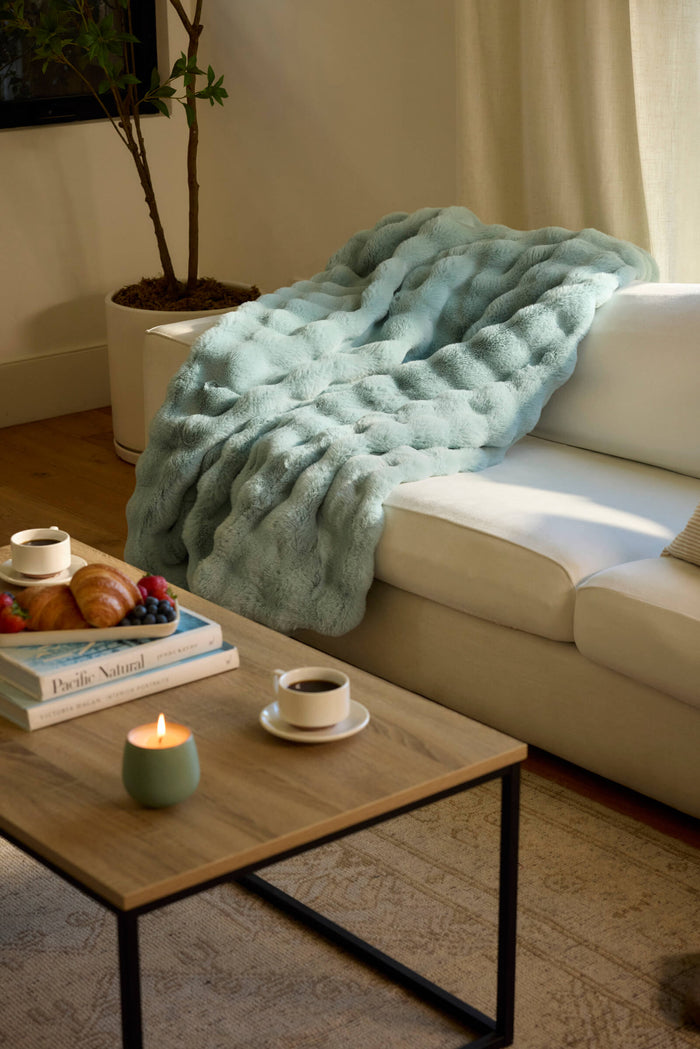 A cozy living room with a white sofa draped in the Cozy Earth Bubble Cuddle Blanket, a wooden coffee table with a candle, two cups of coffee, books, croissants and berries on a plate, and a potted plant in the corner.|Color:Tide