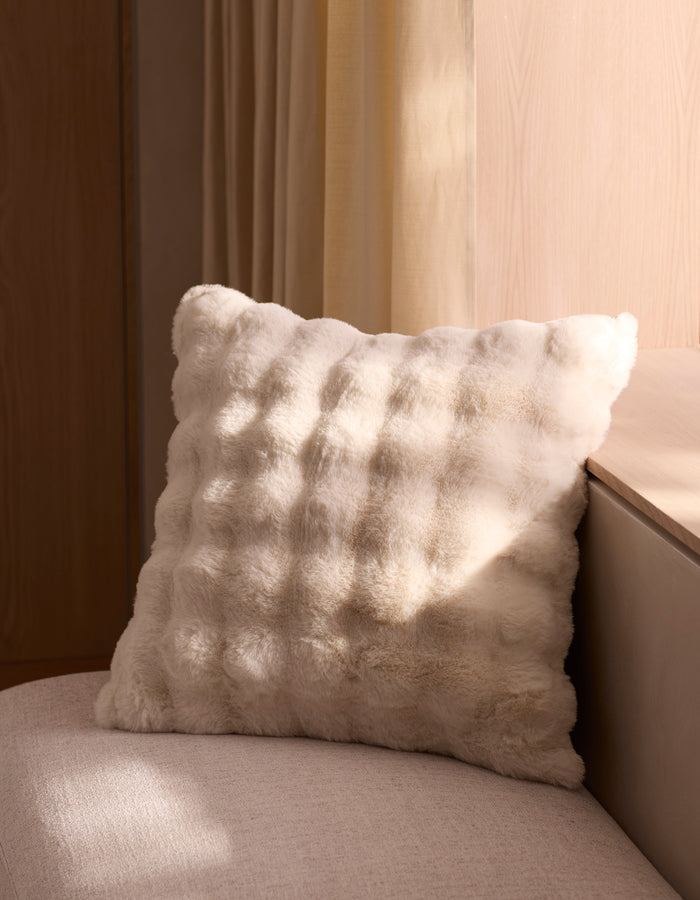 The Cozy Earth Bubble Cuddle Pillow, soft and fluffy in white, sits on a light beige seat near a wall as sunlight streams through the window, casting gentle shadows on the pillow and surrounding surfaces.|Color:Creme |Size:24" x 24"