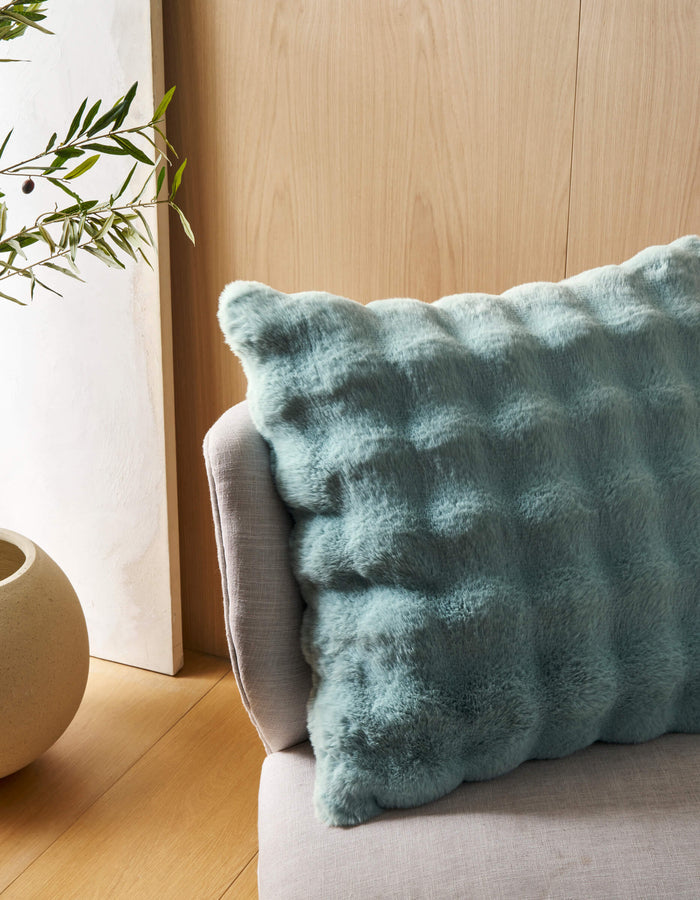 The Bubble Cuddle Pillow by Cozy Earth, featuring a soft textured light blue cover, sits on a light gray upholstered chair beside a round beige planter with greenery, all against a light wood-paneled wall.|Color:Tide |Size:24" x 24"