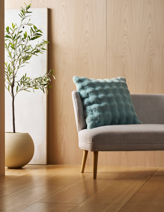 A light gray chair with wooden legs holds a Cozy Earth Bubble Cuddle Pillow in textured blue. Next to it is a potted plant in a beige planter, set before a wooden wall with a minimalist white art piece behind the plant.|Color:Tide |Size:24" x 24"