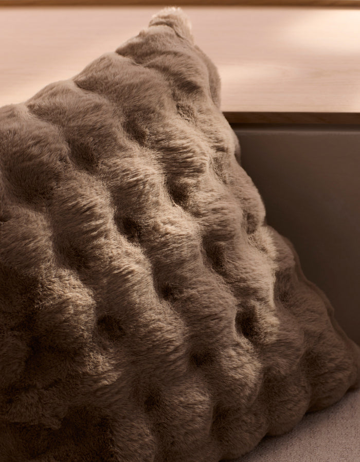 The Cozy Earth Bubble Cuddle Pillow, with its plush brown wavy tufted texture, rests on a light wooden bench in soft natural light.|Color:Walnut |Size:24" x 24"