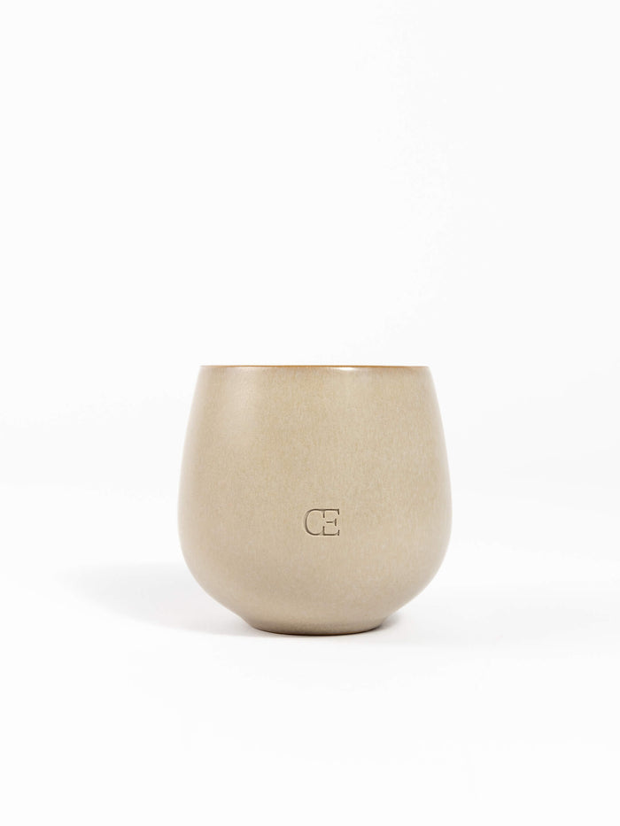 A beige, round ceramic Candle EMBER by Cozy Earth with a matte finish and "CE" embossed on the front, shown against a plain white background.