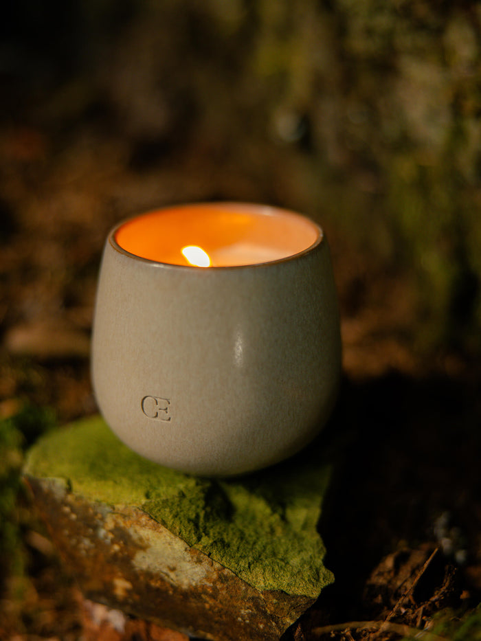 The Candle EMBER by Cozy Earth, in a sleek gray ceramic holder, sits on a mossy rock and casts a warm glow in a serene outdoor setting.