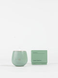 A pale green Cozy Earth Candle UNWIND in a round container, with a small flame, sits beside its matching box labeled "UNWIND" and "Cozy Earth" on a plain white background.