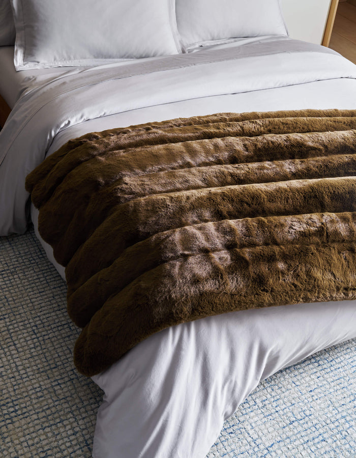 A neatly made bed with white sheets and pillows features the plush Cozy Earth Channel Cuddle Blanket in brown draped across the foot, on top of a blue and white textured rug.|Color:Cedar