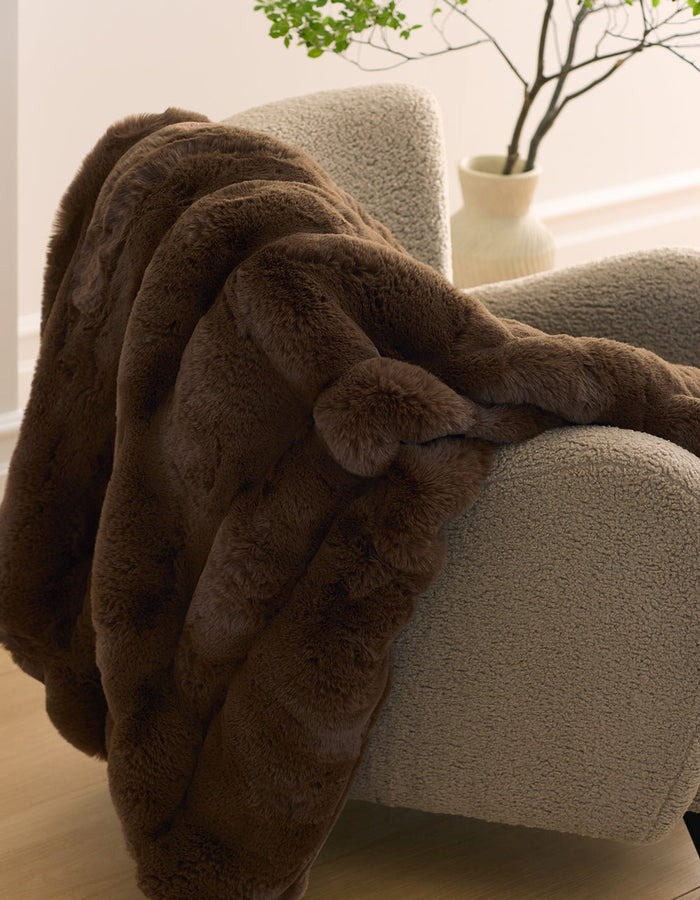 The Cozy Earth Channel Cuddle Blanket, in plush dark brown faux fur, is draped over a textured light beige armchair, with a cream vase of green branches in the background.|Color:Cedar