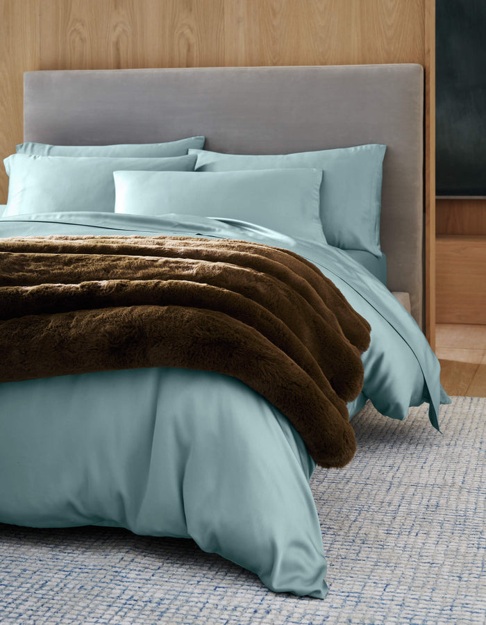 A neatly made bed features a Cozy Earth Channel Cuddle Blanket, light blue bedding, matching pillows, and a soft gray upholstered headboard against a light wood paneled wall on a textured blue rug.|Color:Cedar
