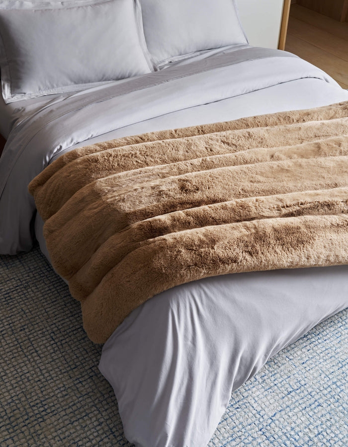 A neatly made bed with white sheets and pillows is topped with the Cozy Earth Channel Cuddle Blanket in thick tan faux fur, set on a textured blue and white rug.|Color:Brandy