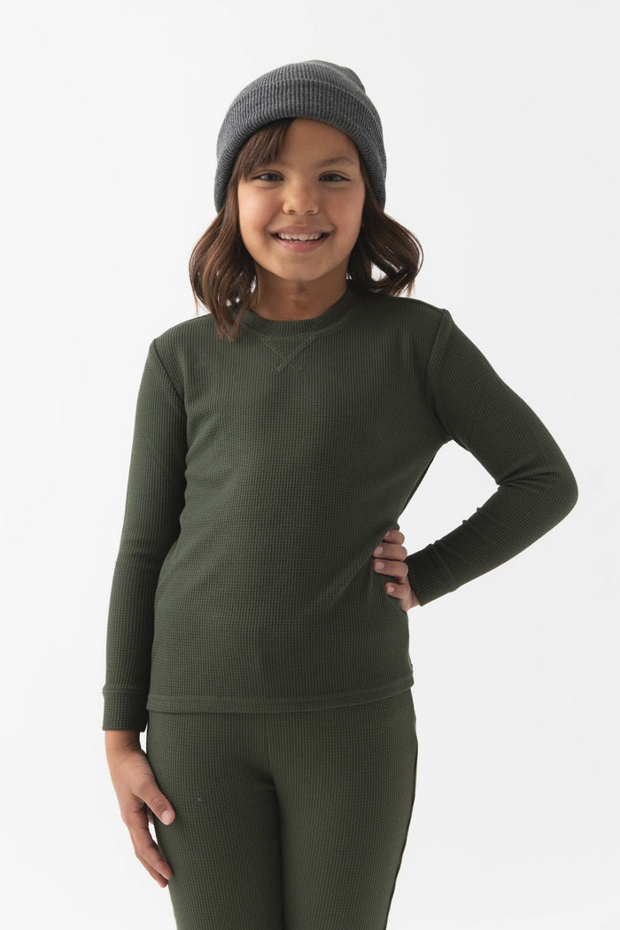 Smiling child with medium-length brown hair, wearing a gray beanie and Cozy Earth Children's Bamboo Waffle Knit Base Layer Long Sleeve in green, standing with one hand on their hip against a plain white background.|Color:Olive