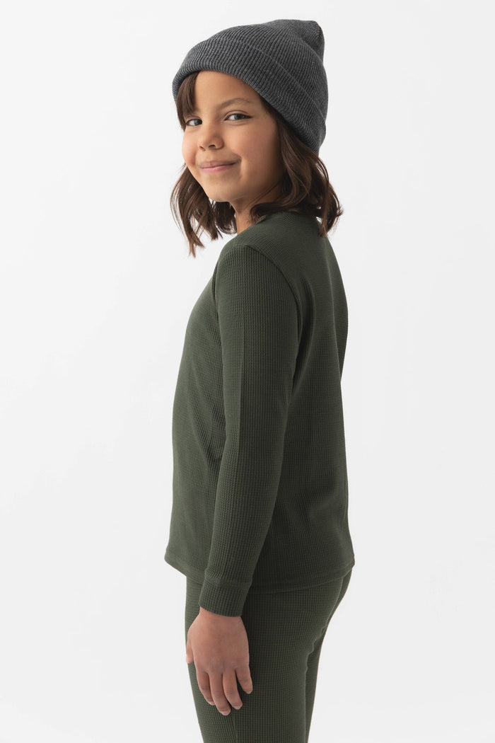A young child stands sideways, smiling at the camera in a Cozy Earth Children's Bamboo Waffle Knit Base Layer Long Sleeve against a plain white background.|Color:Olive