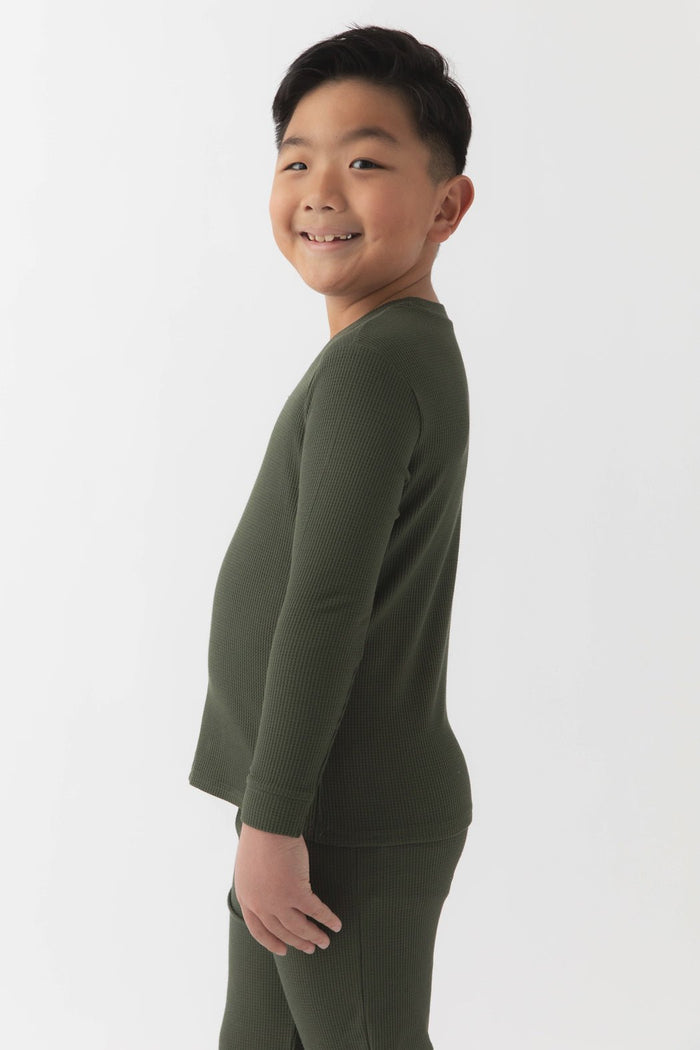 A young boy with short black hair smiles sideways, wearing Cozy Earth's Children's Bamboo Waffle Knit Base Layer Long Sleeve in dark green, paired with matching pants against a plain white background.|Color:Olive