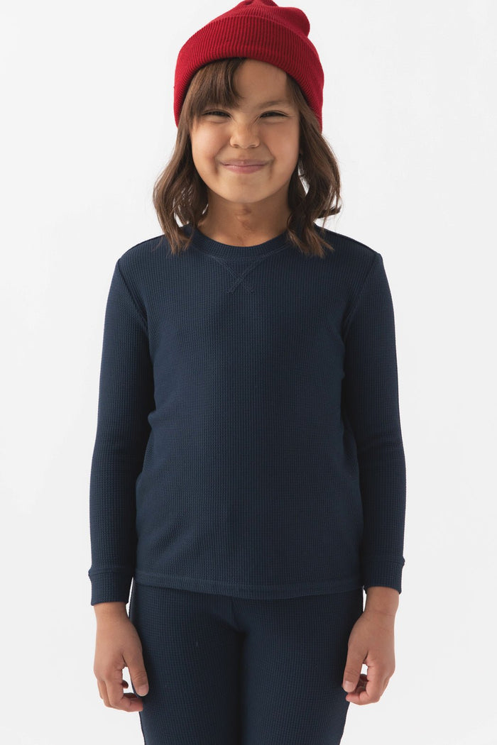 A young child smiles in a red beanie and Cozy Earth's Children's Bamboo Waffle Knit Base Layer Long Sleeve, paired with matching navy blue pants, standing against a plain white background.|Color:Navy