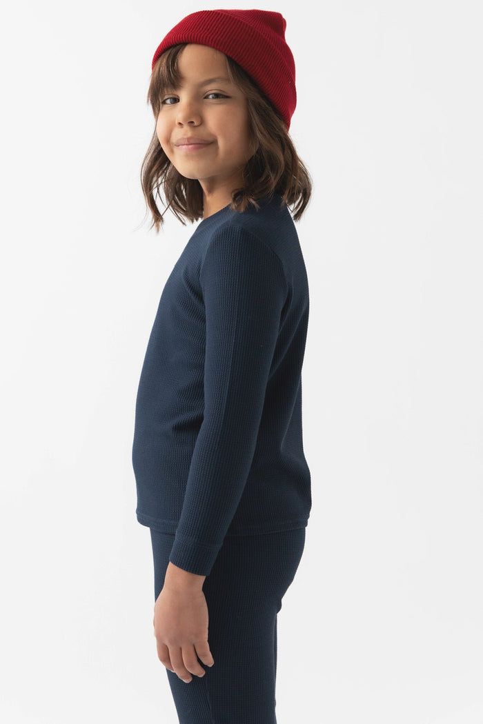 A young child with shoulder-length dark hair wears Cozy Earth's Children's Bamboo Waffle Knit Base Layer Long Sleeve in navy blue, paired with matching pants and a red knit beanie, standing sideways and smiling against a plain white background.|Color:Navy