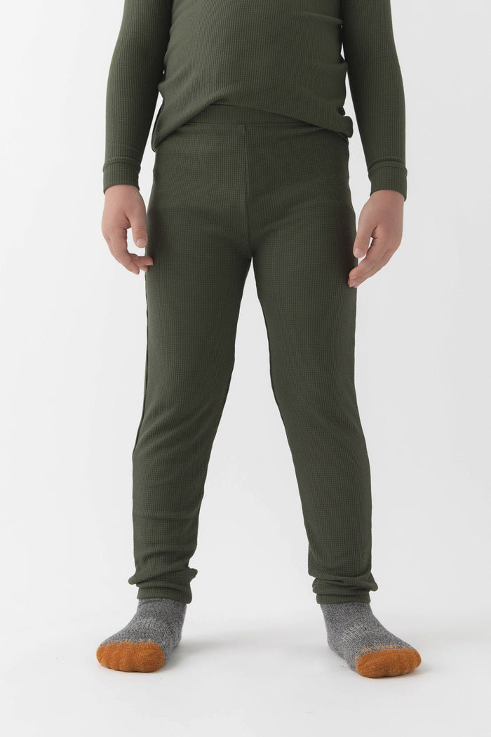 A child wears Cozy Earth’s Children’s Bamboo Waffle Knit Base Layer Pant in Olive, paired with a matching long-sleeve top and gray socks with orange toes, standing against a plain white background. The child’s face is not shown.|Color:Olive