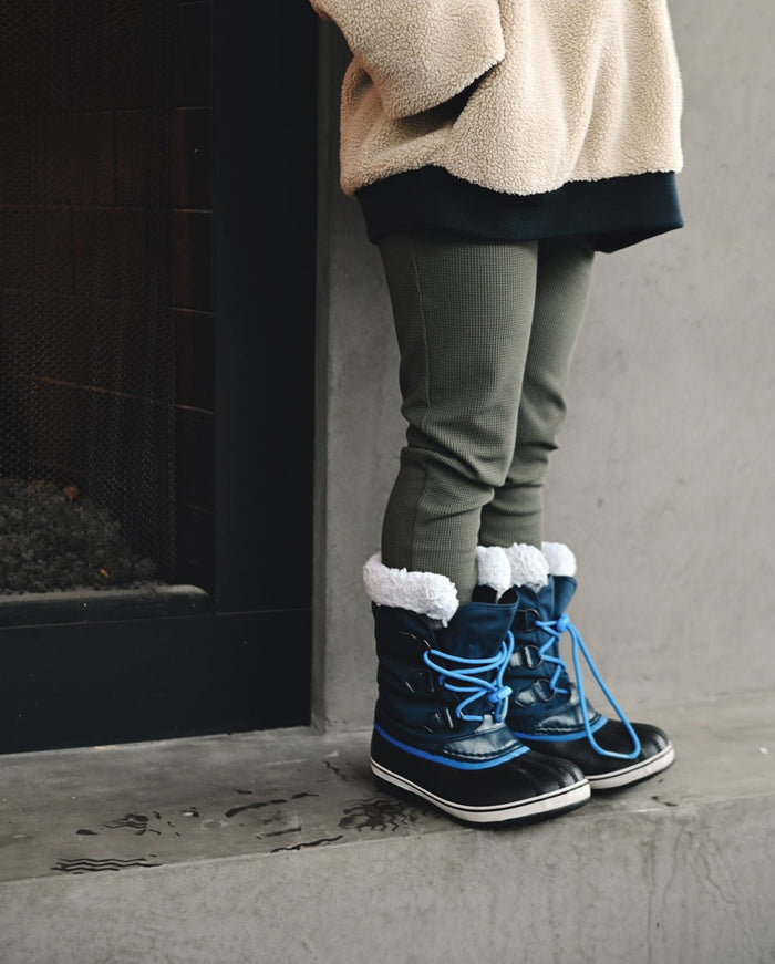 A child in Cozy Earth's Children's Bamboo Waffle Knit Base Layer Pant stands indoors near a fireplace, paired with a beige fleece jacket and blue winter boots with white fur trim on a concrete floor.|Color:Olive