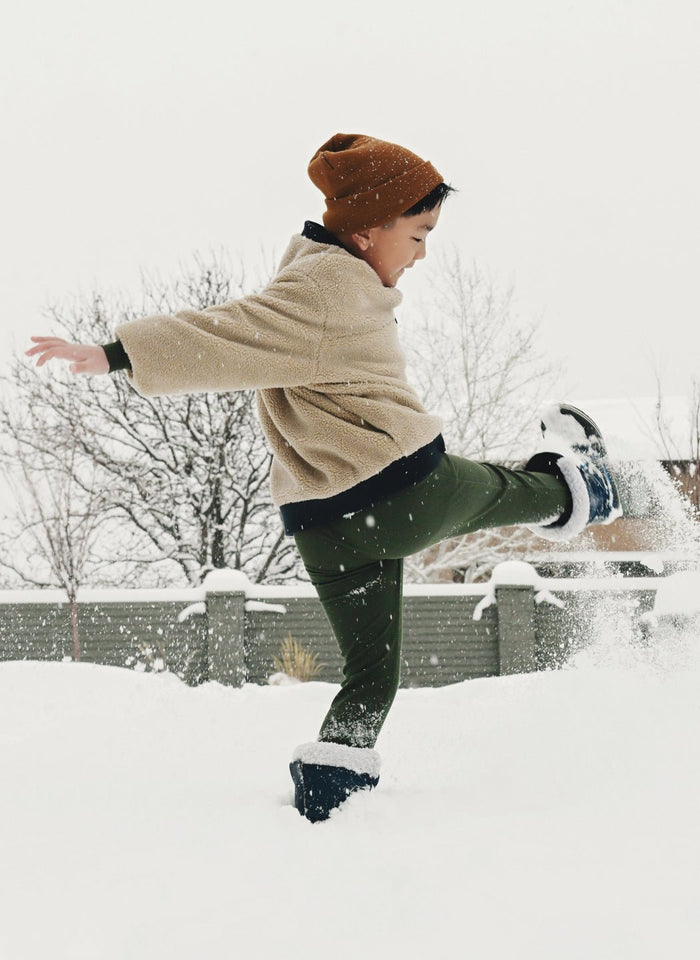 A young child wearing Cozy Earth's Children's Bamboo Waffle Knit Base Layer Pant plays outside, kicking up snow on a wintry day with trees and a fence in the background.|Color:Olive