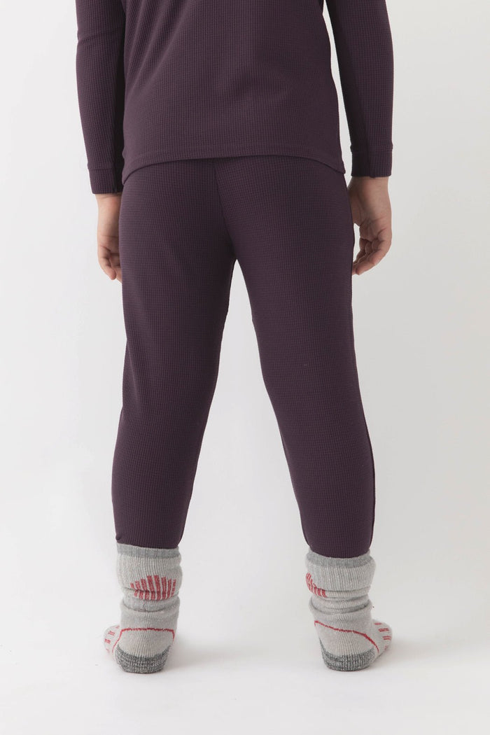 A child faces away, wearing Cozy Earth's dark purple Children's Bamboo Waffle Knit Base Layer Pant with a matching top and thick gray socks with red accents. The background is plain white.|Color:Plum
