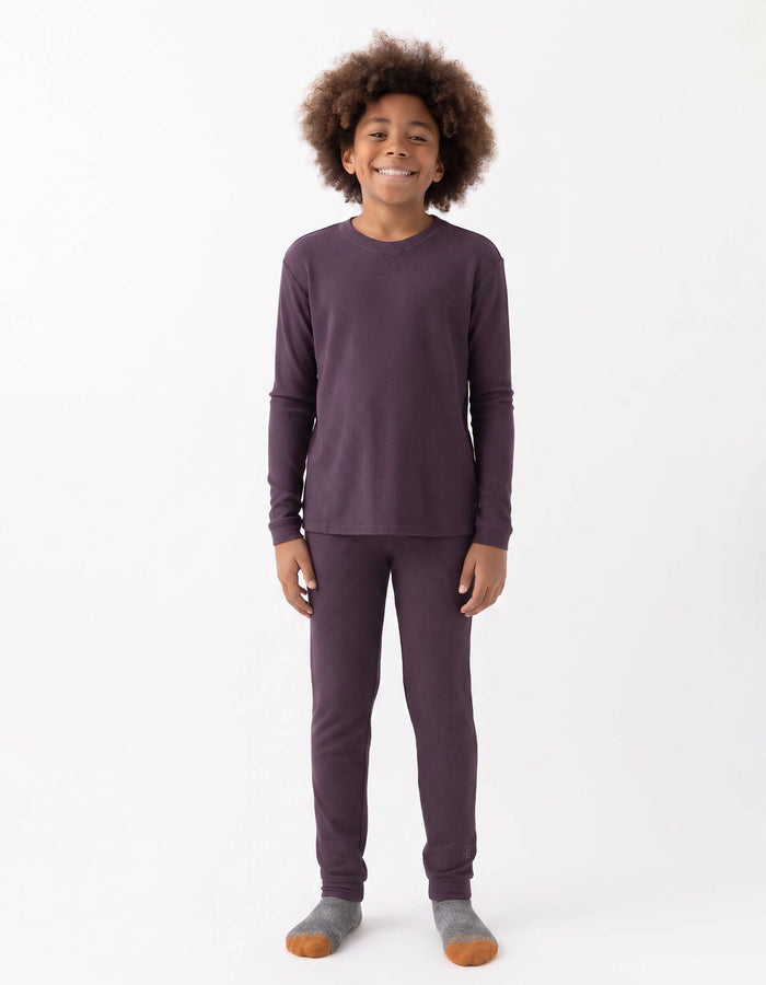 A smiling child with curly hair stands against a white background, wearing the Cozy Earth Children's Bamboo Waffle Knit Base Layer Set - Last Chance, paired with gray and orange socks.|Color:Plum
