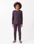 A smiling child with curly hair stands against a white background, wearing the Cozy Earth Children's Bamboo Waffle Knit Base Layer Set - Last Chance, paired with gray and orange socks.|Color:Plum