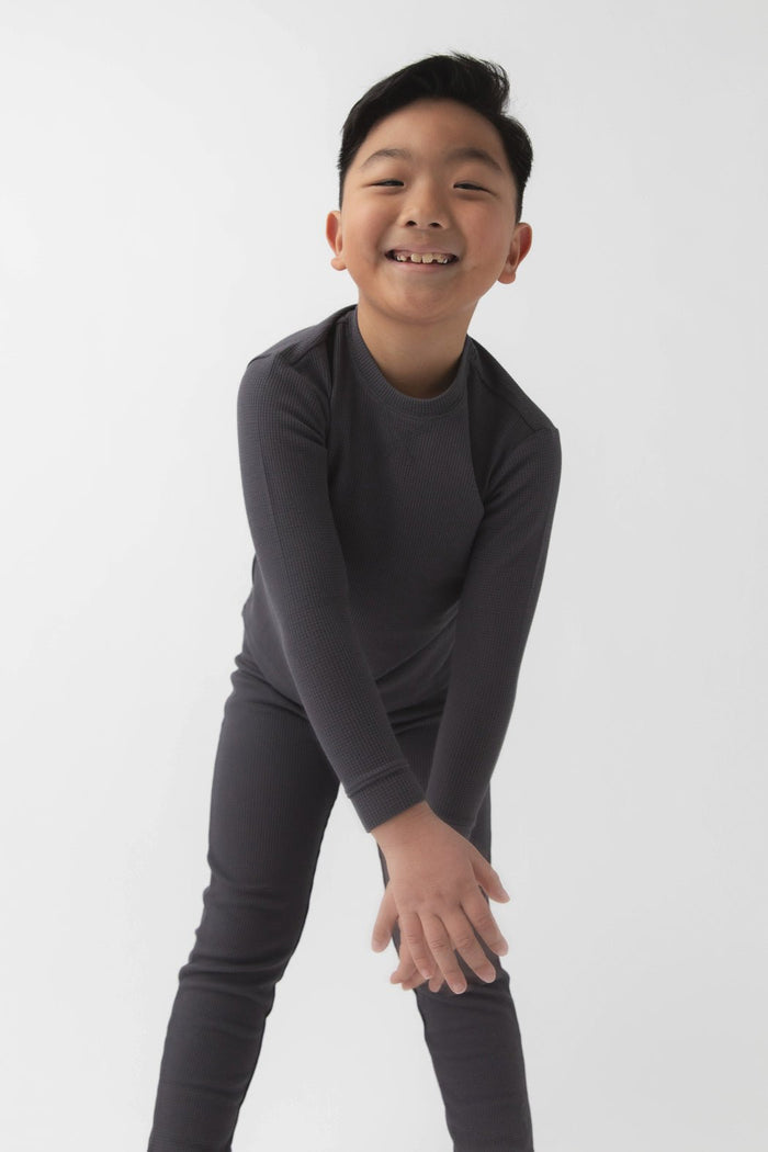 A young boy with short black hair smiles while leaning forward, wearing the Cozy Earth Children's Bamboo Waffle Knit Base Layer Long Sleeve and matching pants against a plain white background.|Color:Iron