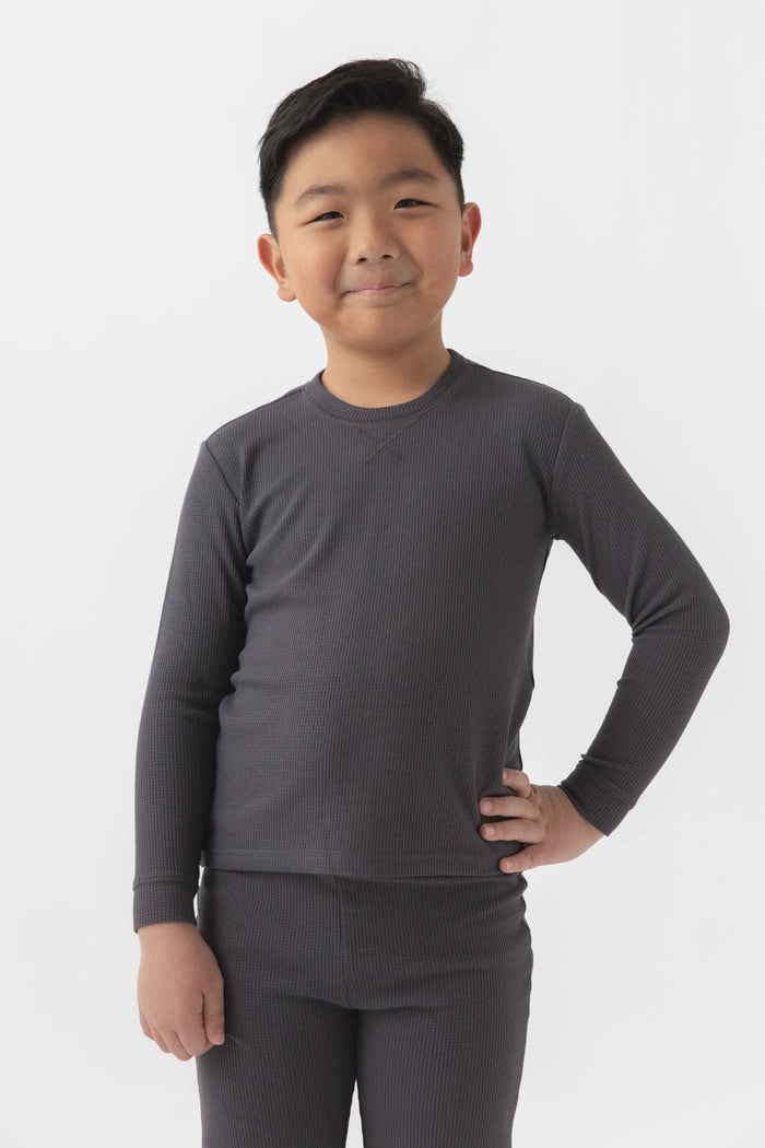 A young boy with short dark hair smiles, standing with one hand on his hip, wearing the Cozy Earth Children's Bamboo Waffle Knit Base Layer Long Sleeve and matching bottoms against a plain white background.|Color:Iron