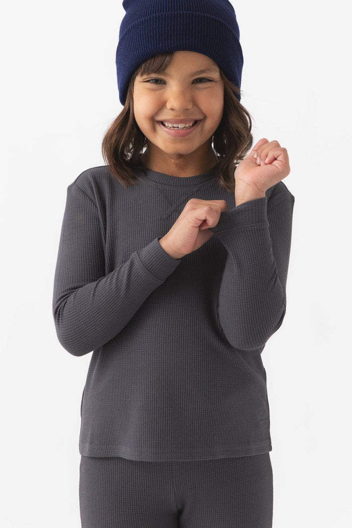 A young child wearing Cozy Earth's Children's Bamboo Waffle Knit Base Layer Long Sleeve in dark gray smiles while holding the sleeve. The beanie is dark blue and the background is plain white.|Color:Iron