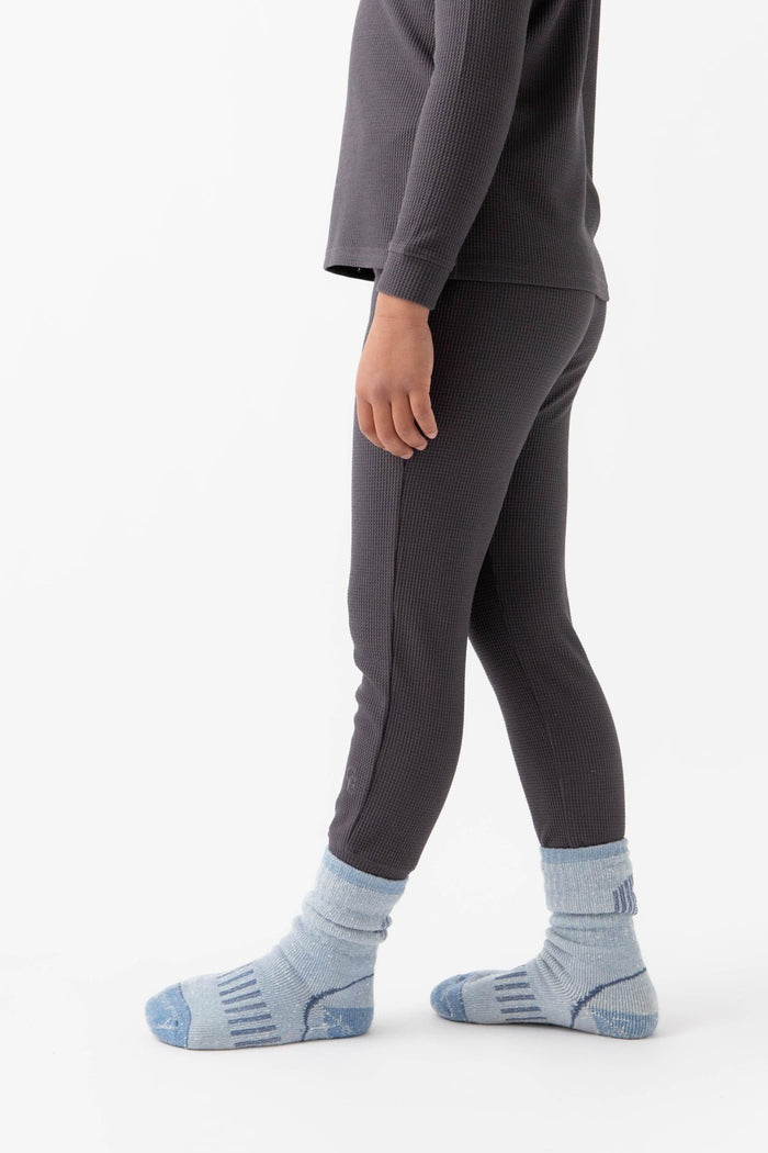 A child wears Cozy Earth's Children's Bamboo Waffle Knit Base Layer Pant in dark gray with a matching ribbed top and light blue socks, standing sideways against a plain white background.|Color:Iron