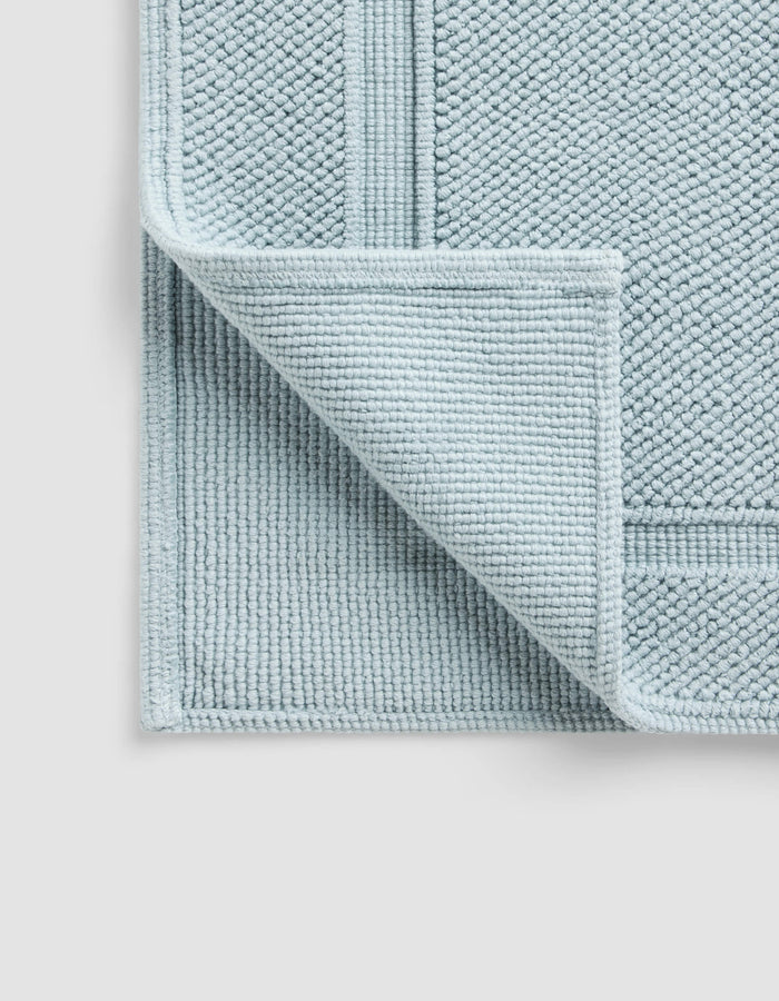 The Cozy Earth Classic Bath Mat, in light blue with a textured surface and border pattern, is shown with one corner flipped to display the underside, resting on a plain, light-colored background.|Color:Harbor Mist
