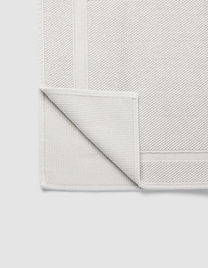 The Cozy Earth Classic Bath Mat in light grey is displayed flat on a plain white background, with one corner folded over to show its woven underside and detailed border.|Color:Light Grey