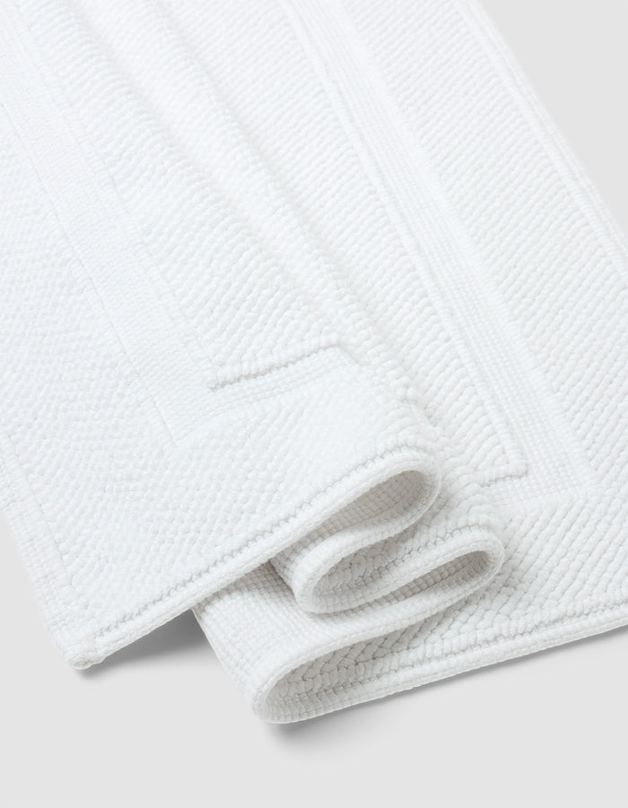 Two neatly folded Cozy Earth Classic Bath Mats, featuring a white textured surface and rectangular border design, are placed on a light gray background.|Color:White