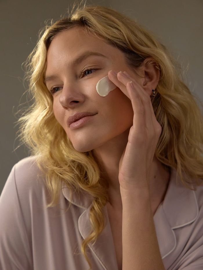 With long, wavy blonde hair, a person applies Cozy Earth's Clay Mask to their cheek, appearing at ease. They're dressed in a pale pink top with a gentle smile. The neutral background emphasizes their skincare ritual. |Size:Full Size
